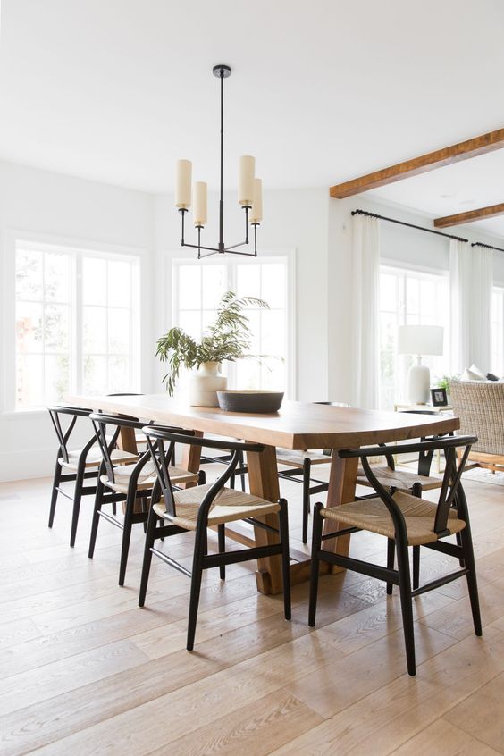 a modern farmhouse dining space with a stained table, black chairs with woven seats, a chandelier, some greenery and a bowl
