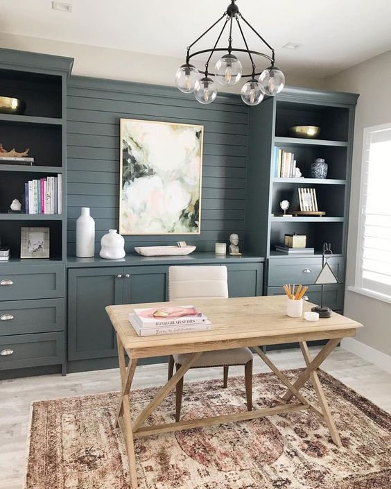 A modern farmhouse home office with a grey built in storage unit, open sheves, a trestle desk and a neutral chair, a chandelier