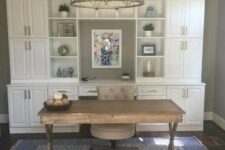 a modern farmhouse home office with a white storage unit, a wooden trestle desk and a comfy chair plus a gold chandelier
