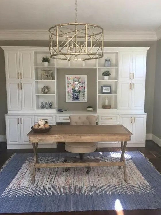 a modern farmhouse home office with a white storage unit, a wooden trestle desk and a comfy chair plus a gold chandelier