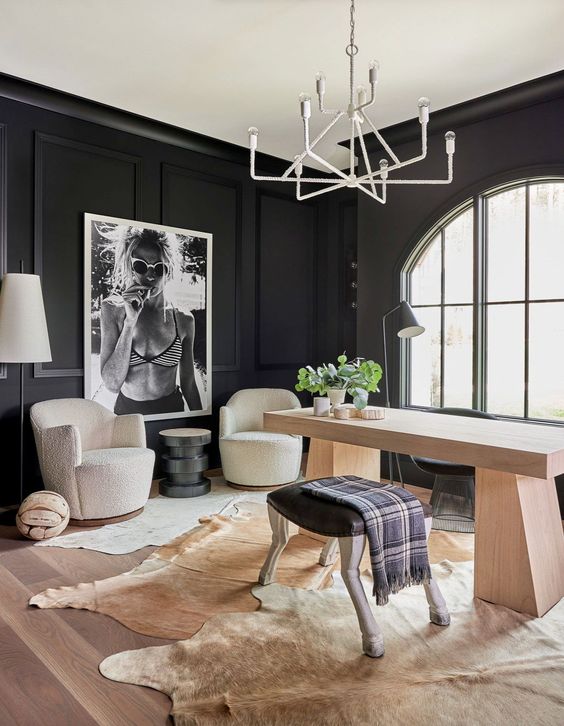 a modern farmhouse home office with black paneled walls, a stained desk, white chairs, a stool, cowhide rugs and a chandelier
