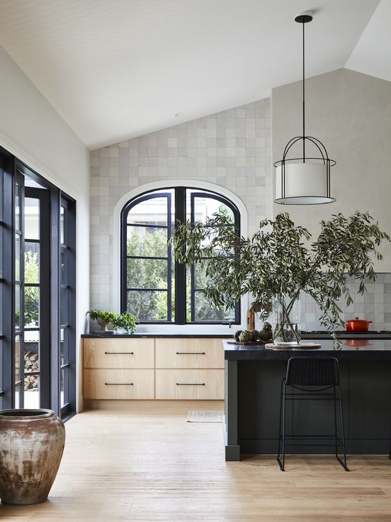 a modern farmhouse kitchen with white Zellige tiles, stained cabinets, a black kitchen island, an arched window and pendant lamps