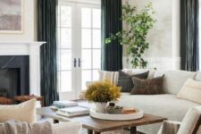 a modern farmhouse living room with a fireplace, creamy sofas and chairs, a stained coffee table, dark curtains and greenery
