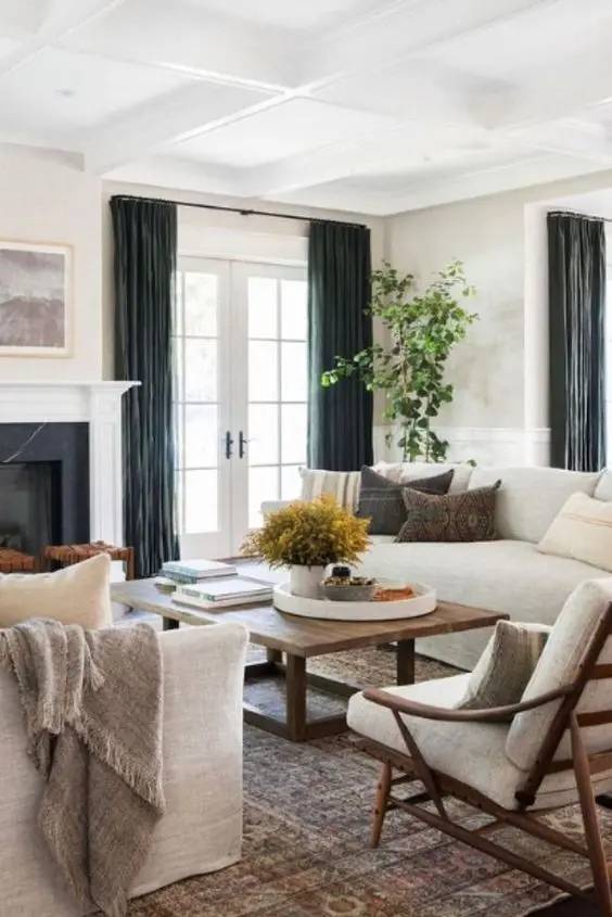 a modern farmhouse living room with a fireplace, creamy sofas and chairs, a stained coffee table, dark curtains and greenery