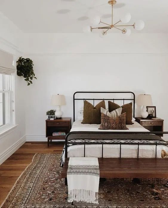 A modern farmhouse meets boho bedroom with a wrought bed and contrasting bedding, a stained bench, a printed rug, dark stained nightstands and potted greenery