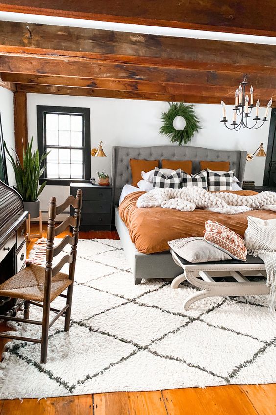 a modern farmhouse meets boho bedroom with stained wooden beams, a grey upholstered bed with bright bedding, black nightstands, a chandelier and greenery