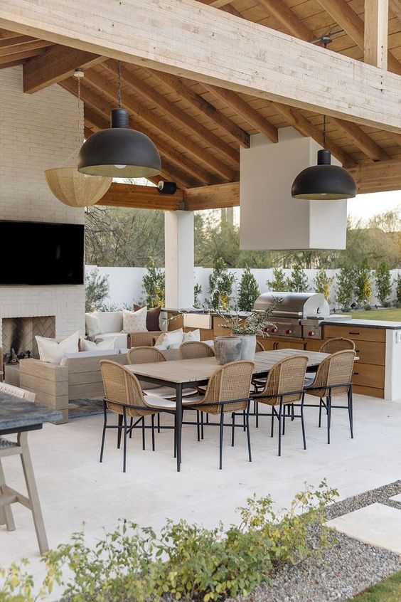 a modern farmhouse terrace with a fireplace, a neutral sofa, a table, wicker chairs, pendant lamps is an inviting and welcoming space