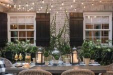 a modern farmhouse terrace with a stained table, wicker chairs, greenery around and on the table, candle lanterns and string lights