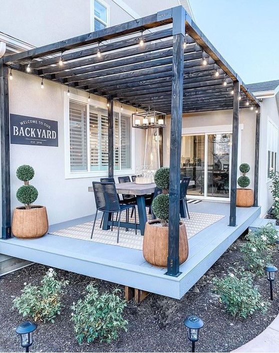 a modern farmhouse terrace with a wood and metal dining table, black metal chairs, potted topiaries and a sign
