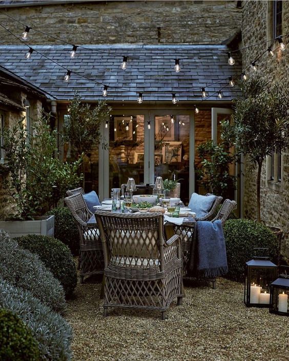 a modern farmhouse terrace with potted trees and greenery, wicker furniture, blue pillows and blankets, string lights and candle lanterns