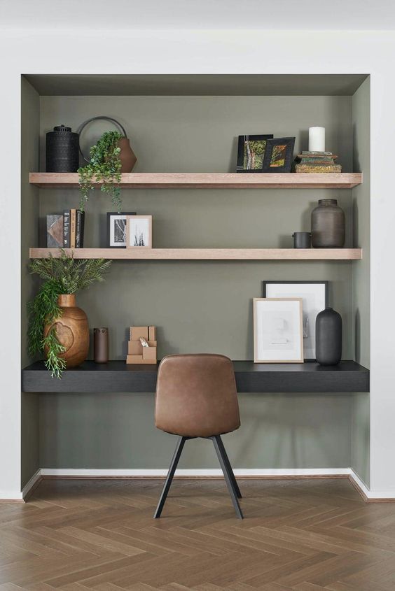 A modern niche painted olive green, with built in shelves and a black desk, some decor and greenery, a brown leather chair and artwork