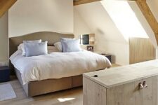 a modern rustic attic bedroom with wooden beams, a neutral upholstered bed, neutral bedding and nneutral planked furniture