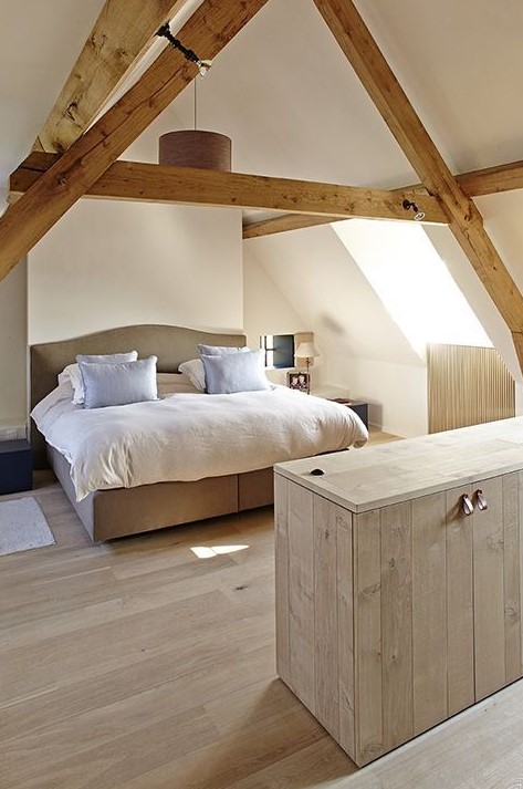 a modern rustic attic bedroom with wooden beams, a neutral upholstered bed, neutral bedding and nneutral planked furniture