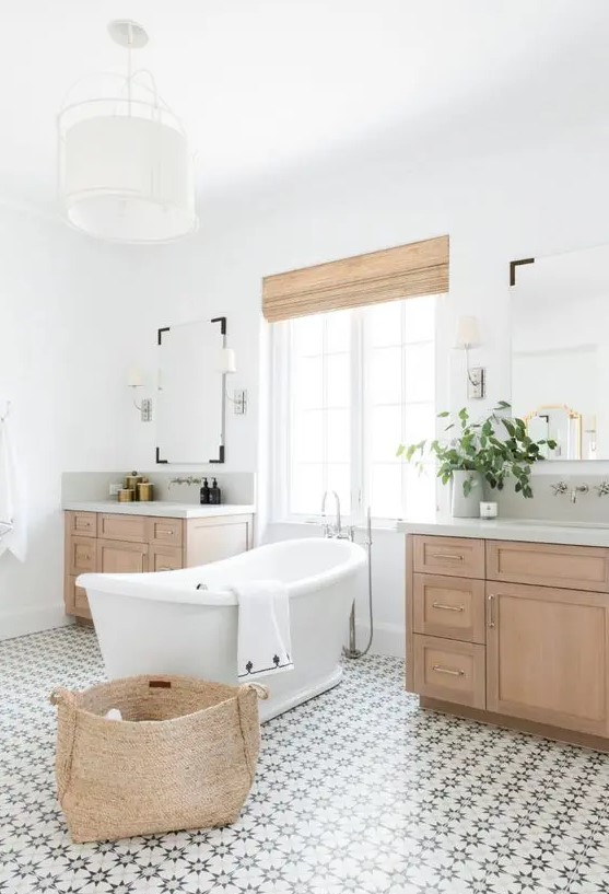 a modern rustic bathroom with a lovely patterned tile floor, stained vanities, a basket and shades plus a vintage-inspired tub