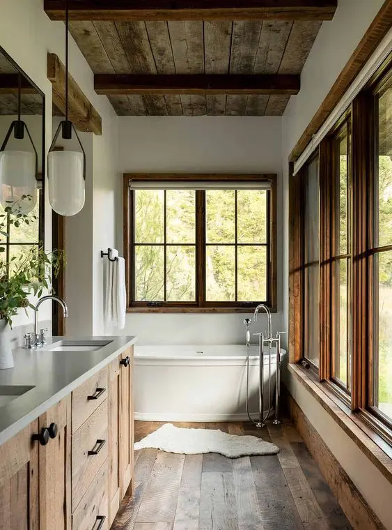 a modern rustic bathroom with lots of windows, a tub, a stained wood vanity, a stained wooden floor, greenery and pendant lamps