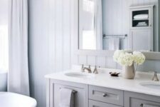 a neutral modern country bathroom with pale blue planked walls, a floating grey vanity, a large mirror, pendant lamps and neutral textiles