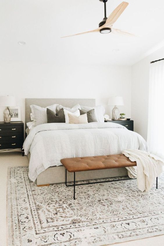 a neutral modern farmhouse bedroom with a grey upholstered bed with neutral bedding, a leather bench, black nightstands that are dressers