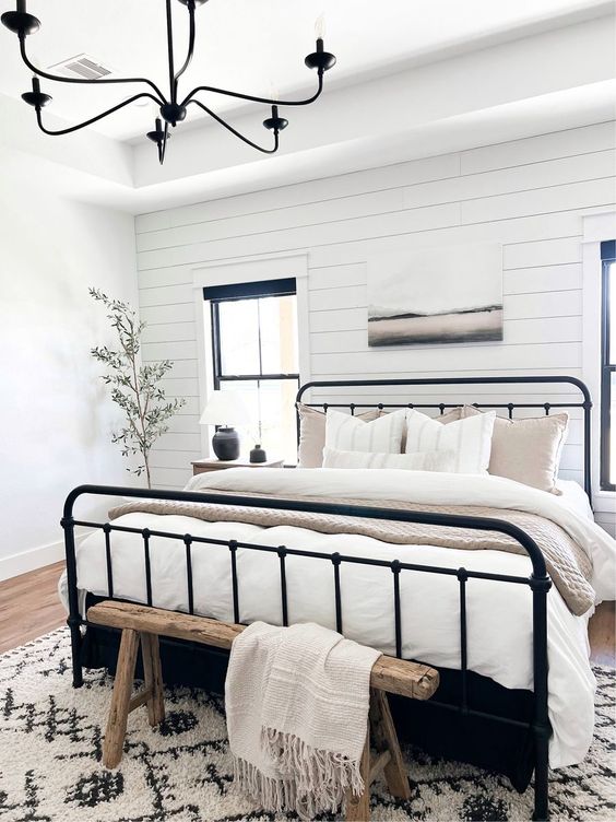 a neutral modern farmhouse bedroom with a shiplap accent wall, a wrought bed with neutral bedding, a stained bench and a printed rug