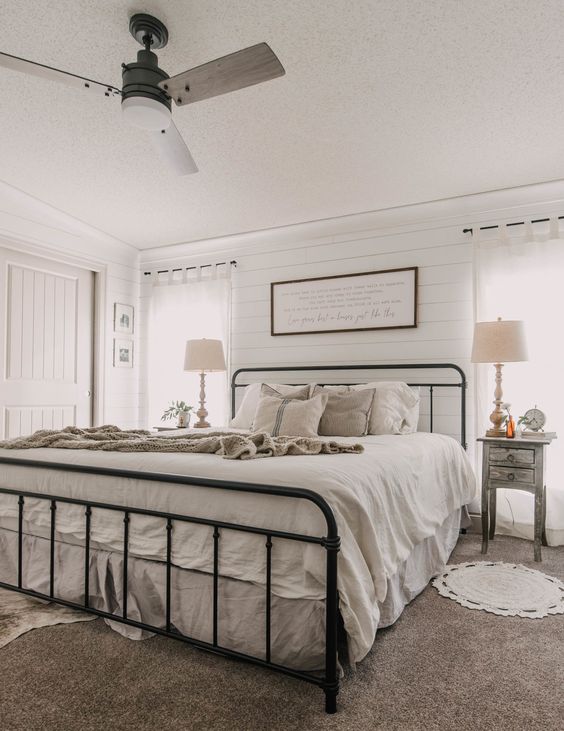 a neutral modern farmhouse bedroom with shiplap walls, a wrought bed with neutral bedding, shabby chic nightstands and some decor