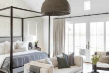 a neutral modern farmhouse bedroom with wooden beams, a blakc frame bed with neutral bedding, creamy seating furniture and a woven pendant lamp
