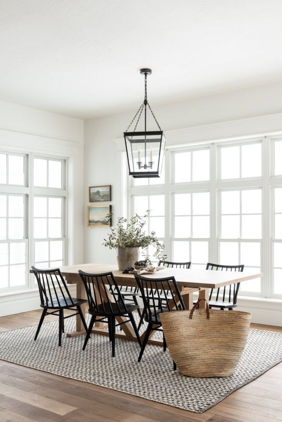 a neutral modern farmhouse dining room with a stained table and black chairs, a black frame pendant lamp, a woven rug and some art and greenery