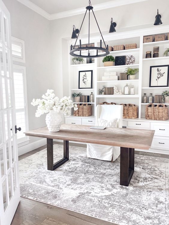 A neutral modern farmhouse home office with a large built in storage unut, a desk, a white chair, a black chandelier and matching sconces