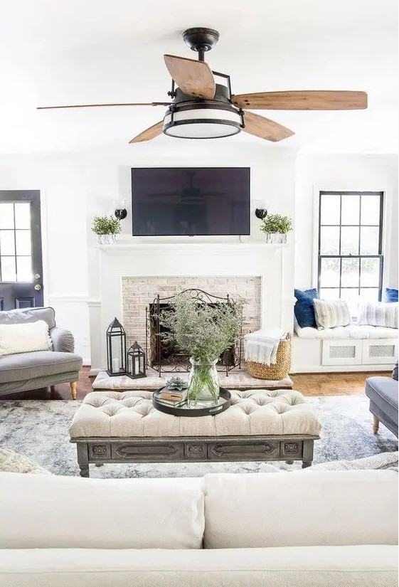 A neutral modern farmhouse living room with a fireplace clad with brick, neutral furniture and built in daybeds, an upholstered ottoman and a fan