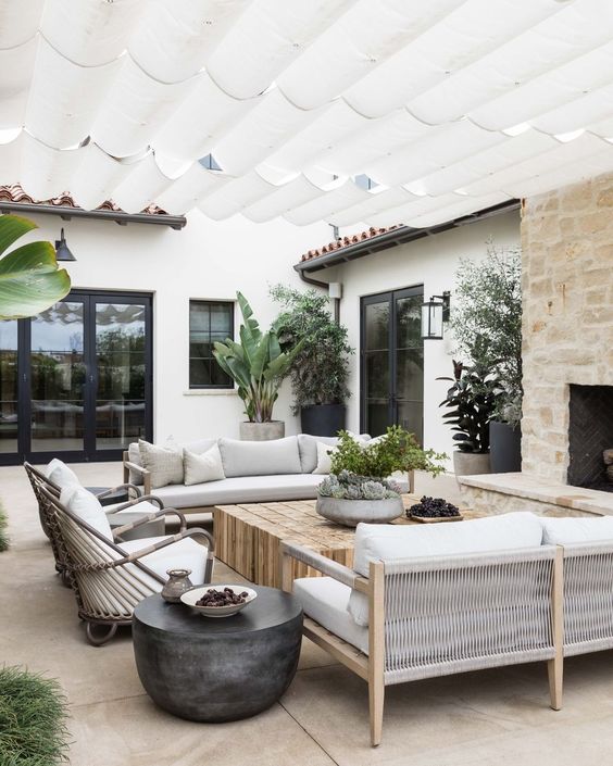 a neutral modern farmhouse terrace with a fireplace, neutral sofas, a low coffee table, rattan chairs, potted trees and greenery