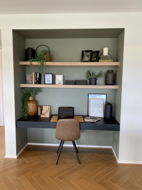 A niche painted olive green, with built in shelves and a black desk, a taupe chair, some decor and potted greenery