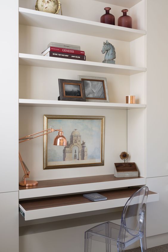 A niche with built in shelves and a desk plus a retracting part, books, artwork, vases and a copper table lamp