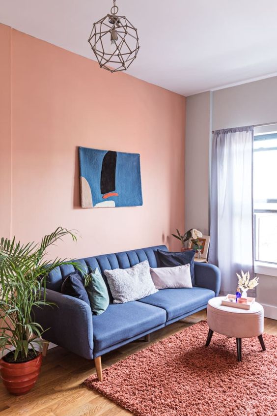 a pink living room with a cobalt blue sofa, a pouf, a coral rug, a blue artwork and potted plants