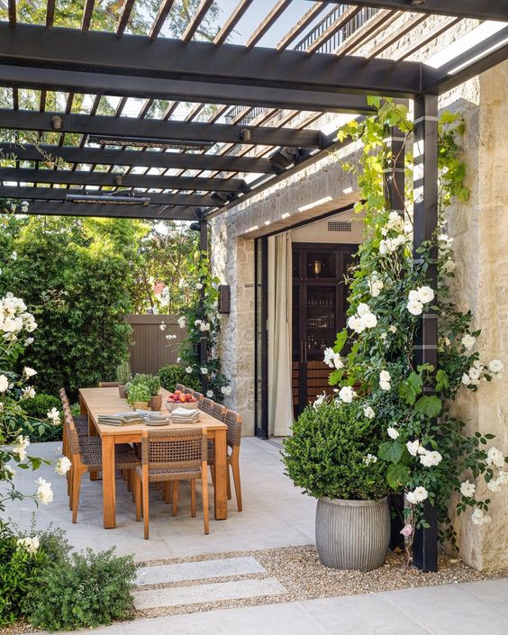 a pretty and welcoming modern farmhouse terrace with vines and white blooms growing, a rustic table and chairs done with cane is amazing