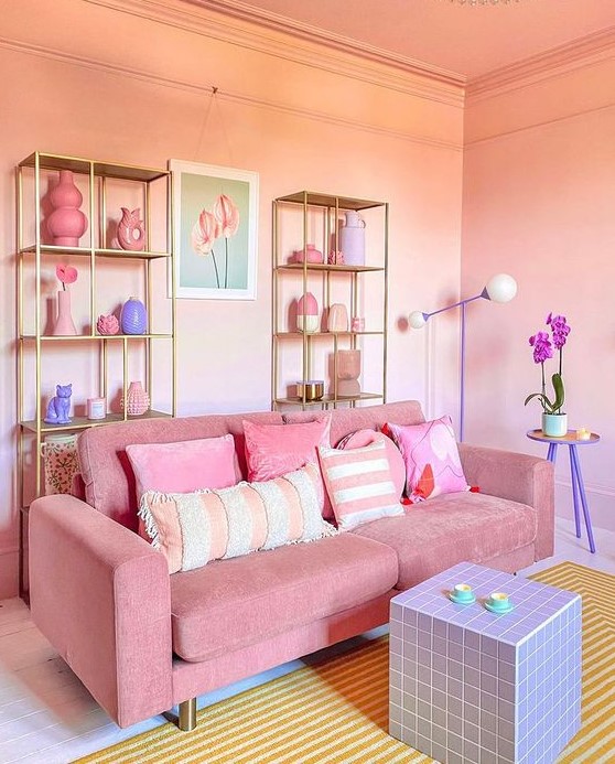 a pretty pink living room with gold shelving units, a pink sofa with pastel pillows, a lilac cube table and a yellow rug