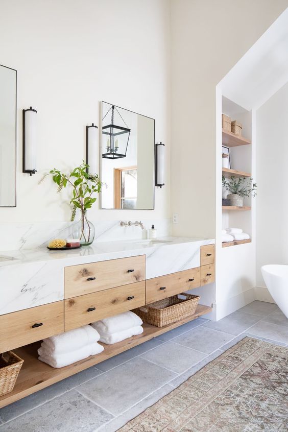 A refined modern farmhouse bathroom with an oval tub, built in shelves, a double timber vanity with a marble countertop, mirrors and sconces