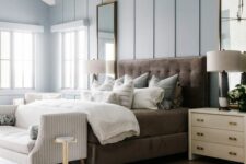 a refined modern farmhouse bedroom with pale blue walls, a brown upholstered bed with neutral bedding, creamy nightstands, a striped bench and a chandelier