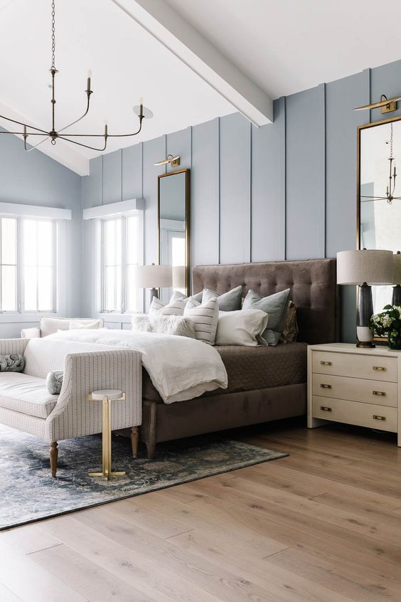 a refined modern farmhouse bedroom with pale blue walls, a brown upholstered bed with neutral bedding, creamy nightstands, a striped bench and a chandelier