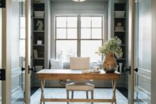 a refined modern farmhouse home office with grey storage units and paneling, a stained desk and a neutral chair, a chic gilded chandelier and greenery
