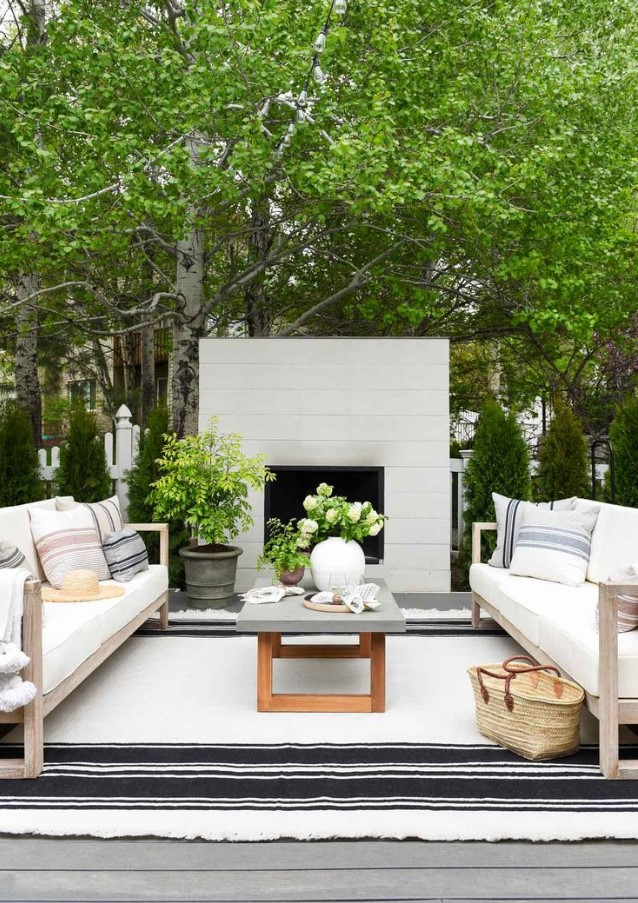 a relaxed modern farmhouse outdoor living room with a fireplace clad with neutral wood, white sofas, a coffee table with a concrete tabletop and striped textiles