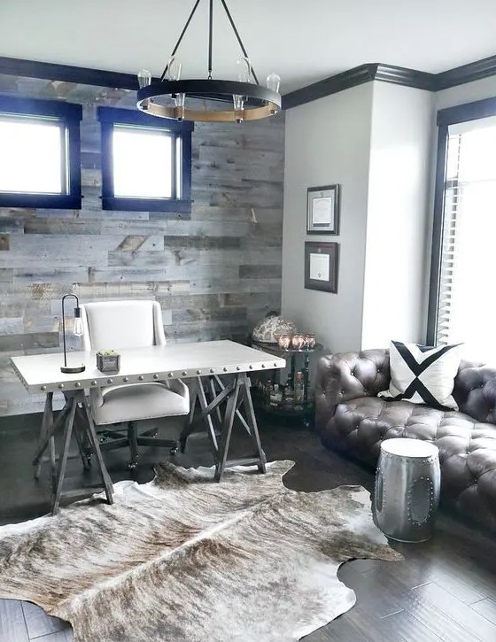 a rustic home office with a salvaged wood wall, a vintage chandelier, a faux fur rug, a trestle desk, a white chair and a leather sofa