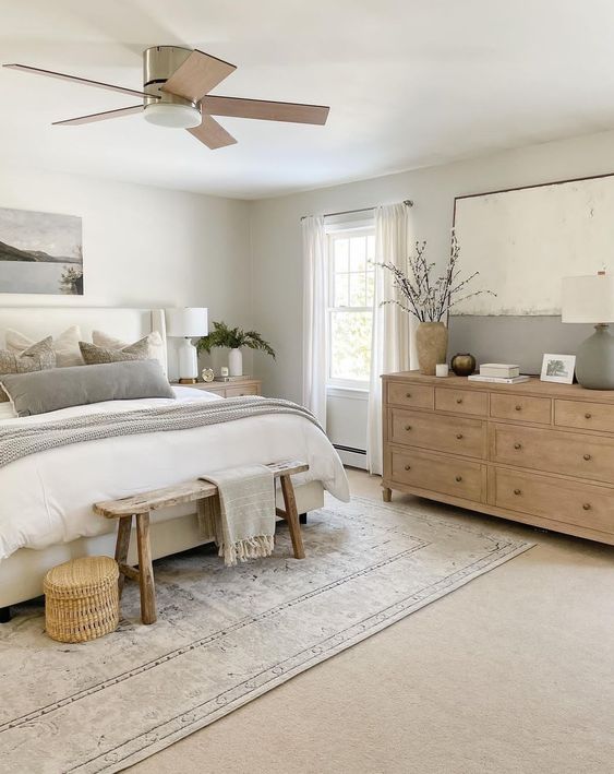 a serene modern farmhouse bedroom with an upholstered bed and neutral bedding, a stained dresser, a stained bench, some decor and a basket