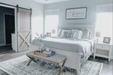 a serene modern farmhouse bedroom with light blue walls, a white bed with grey bedding, white nightstands, a bench and a chic chandelier