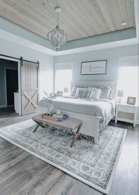 a serene modern farmhouse bedroom with light blue walls, a white bed with grey bedding, white nightstands, a bench and a chic chandelier