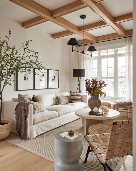 a serene modern farmhouse living room with a coffered ceiling, a creamy sofa and pillows, stools, a woven chair, a coffee table and a black chandelier
