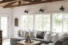 a simple barn living room with white planked walls, a grey sofa, wooden beams, a wooden table and lots of natural light