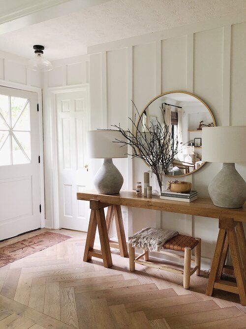 a simple modern farmhouse entryway with a trestle console table, a woven stool, a round mirror, a table lamp