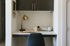 a small and chic home office nook with a built-in cabinet, a built-in desk, a navy chair, a brass lamp and a mirror