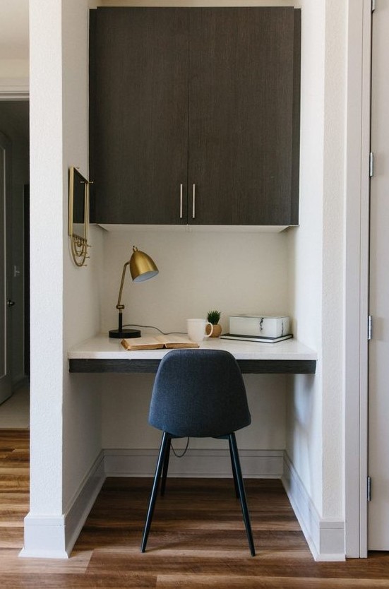 A small and chic home office nook with a built in cabinet, a built in desk, a navy chair, a brass lamp and a mirror