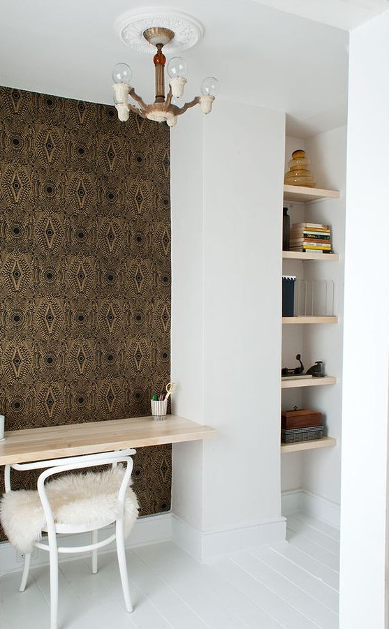 A small home office nook with a series of built in shelves, a desk and a white chair, an accent wall done with wallpaper