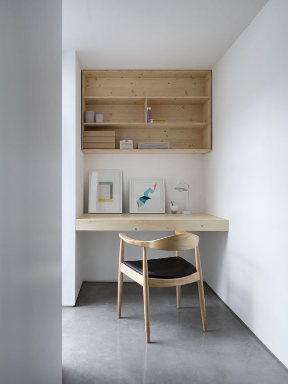 a small minimal niche with a built-in box shelf and a desk, a black chair and some lovely decor, a table lamp