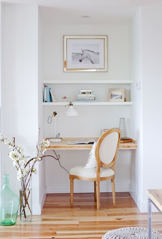 a small nook with built-in shelves and a desk, a white chair, an artwork and a pretty lamp is comfy for working